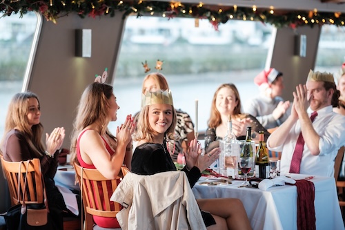 Corporate guests wearing paper crowns and celebrating around a festive dining table on a private Thames boat charter